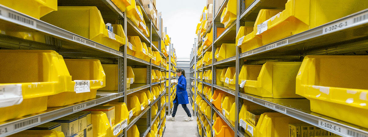 A person in a blue uniform walks through a spacious warehouse, surrounded by shelves and various stored items.